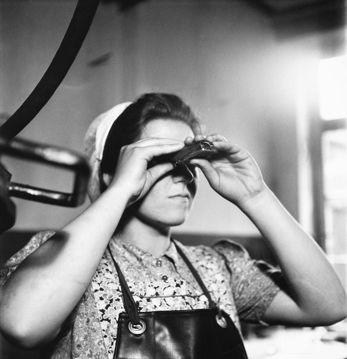 Schwarz-weiß-Fotografie einer jungen Frau in Arbeitskleidung, die konzentriert durch ein kleines optisches Prüfgerät blickt. Sie trägt ein gemustertes Kleid mit Schürze aus Leder und hat ein Tuch über dem Kopf gebunden. Die Szene wirkt wie aus einem handwerklichen oder industriellen Arbeitsumfeld der Mitte des 20. Jahrhunderts. Im Hintergrund fällt Licht durch ein Fenster und erzeugt eine helle Atmosphäre im Raum. Links im Bild sind verschwommene Maschinenteile zu erkennen.