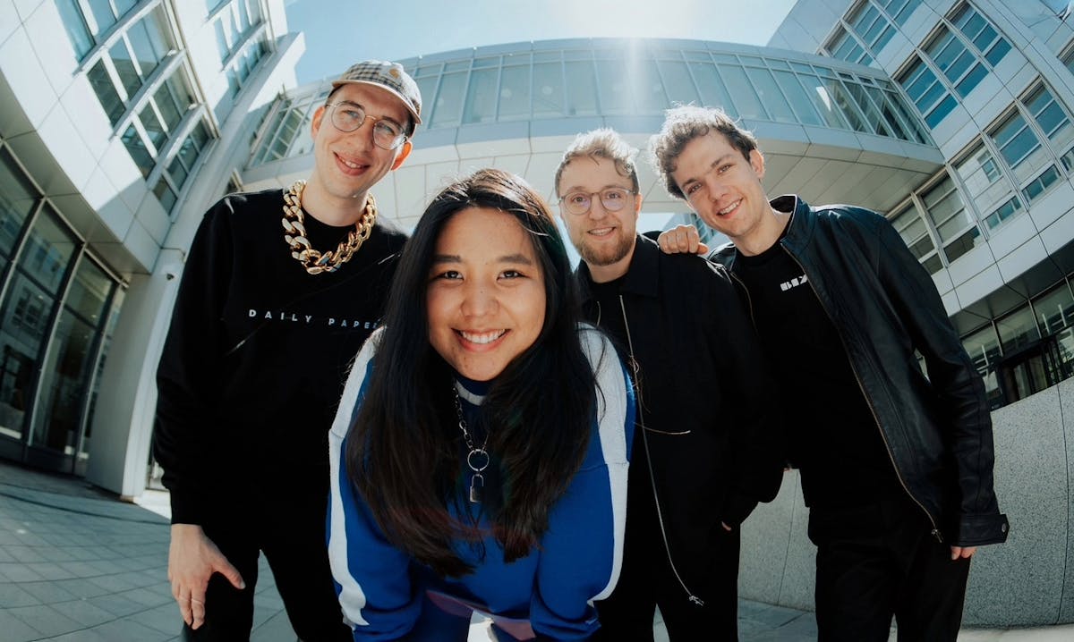 Vier junge Erwachsene stehen lachend dicht beieinander vor einem modernen, verglasten Gebäude. Die Frau in der Mitte trägt einen blauen Pullover und beugt sich freundlich lächelnd zur Kamera. Links von ihr steht ein junger Mann mit Brille, Basecap, schwarzem Pullover und goldener Kette. Rechts von ihr stehen zwei weitere Männer, einer mit Brille und Bart, der andere mit lockigen Haaren und schwarzer Lederjacke. Die Szene wirkt freundlich, urban und dynamisch, aufgenommen bei Sonnenschein aus einer leicht verzerrten Perspektive von unten.