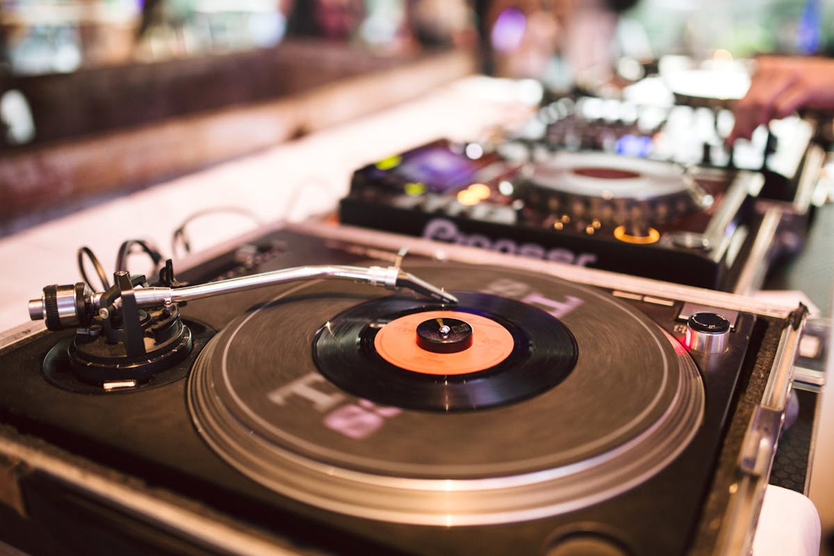 Nahaufnahme eines DJ-Setups mit einem Schallplattenspieler im Vordergrund, auf dem eine schwarze Vinylsingle mit rotem Etikett liegt. Der Tonarm ist über der Platte positioniert. Im Hintergrund sind weitere DJ-Geräte mit beleuchteten Knöpfen und ein Teil einer Hand zu sehen, die offenbar an einem Regler arbeitet. Die Szene wirkt dynamisch und modern, mit leicht verschwommenem Hintergrund.