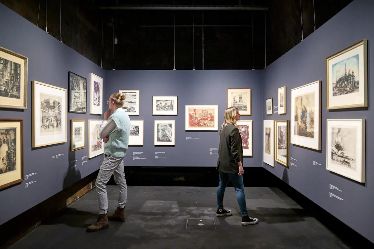 Zwei Personen betrachten gerahmte Kunstwerke in einer Ausstellung mit dunkelblauen Wänden. Die Werke zeigen überwiegend industrielle Motive in unterschiedlichen Zeichen- und Drucktechniken. Links steht eine Person mit zusammengebundenen Haaren und hellblauer Weste nachdenklich vor einem Bild, während rechts eine andere Person mit offenen Haaren und schwarzem Blazer interessiert ein anderes Bild betrachtet. Die Ausstellung ist ruhig und gut beleuchtet.