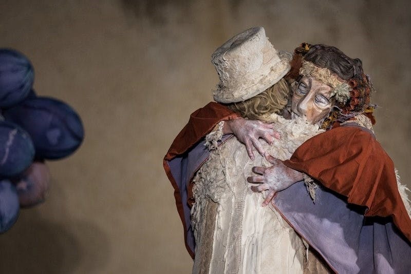 Zwei kunstvoll geschminkte Personen in fantasievollen, historischen Kostümen umarmen sich innig auf einer Bühne. Eine der Figuren trägt einen roten Umhang und eine Blumenkopfbedeckung, während die andere eine helle, abgenutzte Kleidung mit passendem Zylinderhut trägt. Beide wirken wie aus einem Theaterstück oder einer Pantomimendarstellung. Die Szene strahlt emotionale Nähe und Melancholie aus. Links am Bildrand sind unscharfe, lilafarbene Requisiten zu erkennen. Der Hintergrund ist neutral und lenkt den Fokus auf die innige Umarmung der beiden Darsteller.