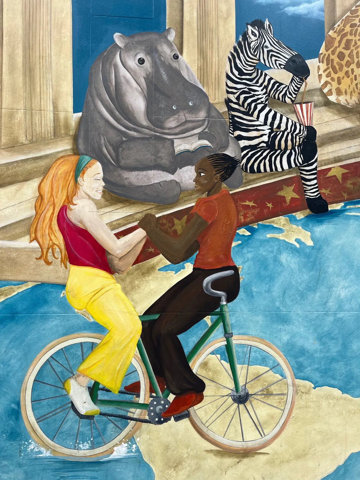 Wandgemälde mit zwei Personen, die gemeinsam auf einem Tandemfahrrad fahren, das über eine Weltkarte rollt. Die Person vorne hat dunkle Haut und kurze Zöpfe, die hintere Person hat helle Haut und langes rotes Haar mit einem türkisfarbenen Haarband. Sie schauen sich an und halten sich an den Händen. Im Hintergrund sitzen auf steinernen Stufen ein lesendes Nilpferd, ein Zebra mit Trinkbecher und Strohhalm sowie ein Teil eines Giraffenkörpers. Die Szene kombiniert reale und phantasievolle Elemente in kräftigen Farben.