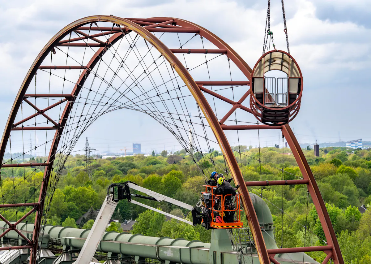 Das Bild zeigt den Abbau des Riesenrads auf dem Gelände des Welterbes Zollverein. Arbeiter in Sicherheitsausrüstung befinden sich in einem Hubkorb und demontieren Teile des Stahlgerüsts. Ein rundes Kabinenteil wird mit einem Kran abgehoben. Die bogenförmige Struktur mit gespannten Seilen gehört zur Konstruktion des Riesenrads. Im Hintergrund ist die grüne Umgebung des Ruhrgebiets mit Industrieanlagen und Strommasten zu sehen.