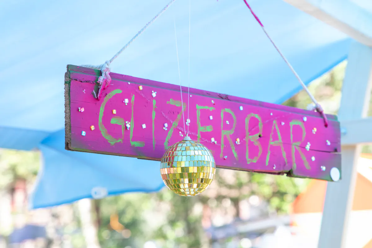 Ein handgemaltes Holzschild mit dem Schriftzug Glitzerbar hängt an zwei weißen Seilen unter einem blauen Sonnensegel. Das Schild ist pink bemalt, mit grüner Schrift und verziert mit kleinen, glitzernden Schmucksteinen. Darunter hängt eine kleine goldene Discokugel, die im Sonnenlicht glänzt. Im Hintergrund sind verschwommen Bäume und weitere Sonnensegel zu erkennen.
