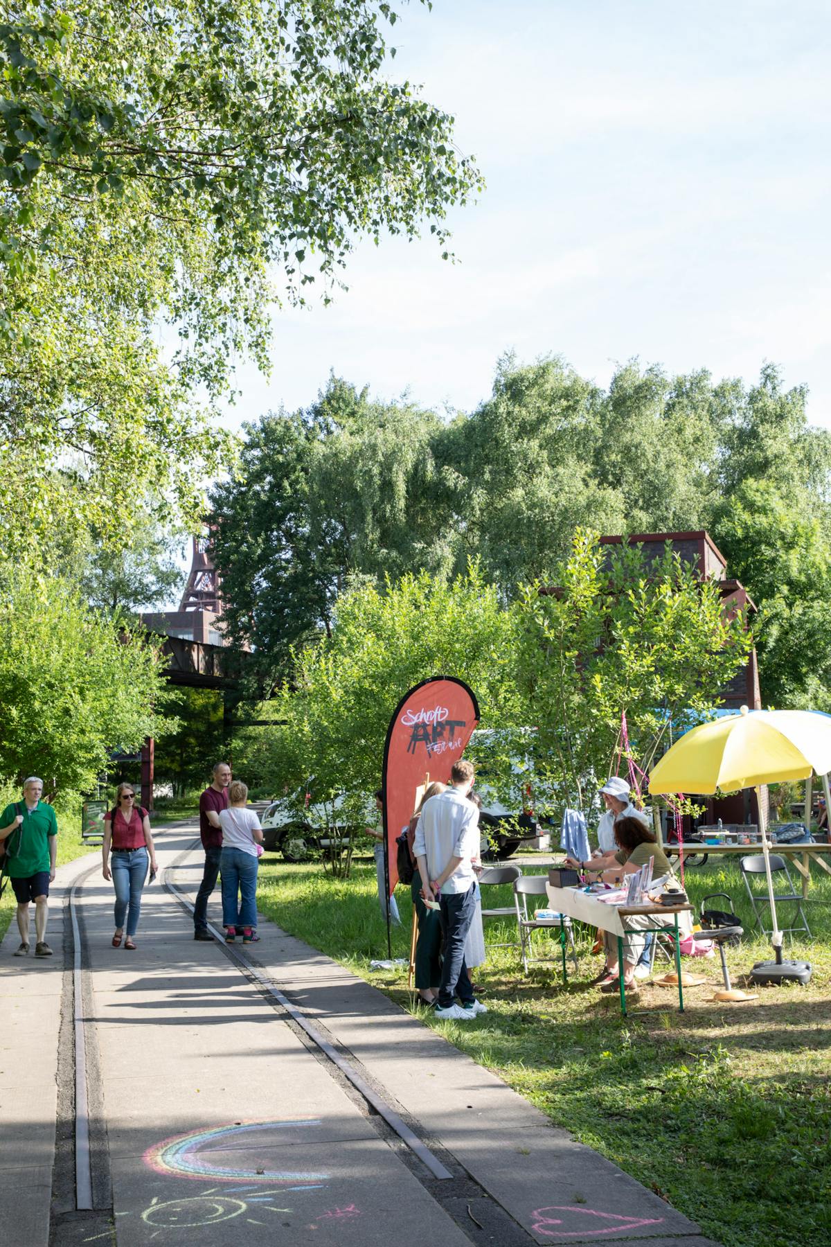 Menschen spazieren bei sonnigem Wetter über einen Weg im Grünen, der von alten Schienen durchzogen ist. Links und rechts säumen Bäume die Szene, im Hintergrund ist ein Förderturm der Zeche Zollverein sichtbar. Auf dem Gehweg ist mit bunter Kreide eine Sonne mit Regenbogen gemalt. Rechts steht ein Info- oder Bastelstand mit gelbem Sonnenschirm und einem roten Banner mit der Aufschrift Schicht ART Festival. Mehrere Personen unterhalten sich oder informieren sich am Stand.
