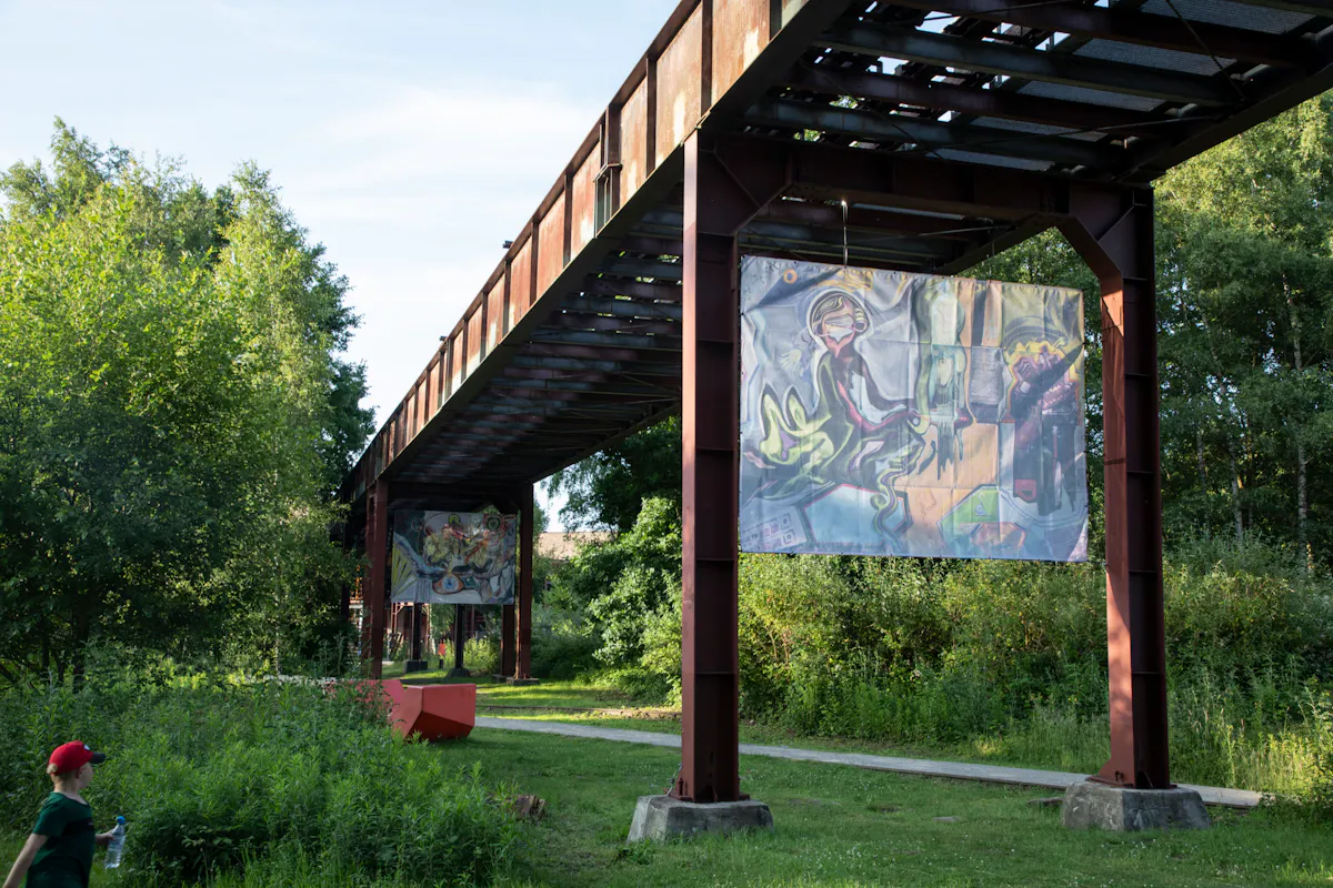 Ein Kind mit roter Kappe läuft an einem sonnigen Tag durch eine grüne Parklandschaft. Über einem Weg verlaufen rostige Stahlträger einer alten Industrieanlage, an denen große, bunte Kunstbanner hängen. Die Banner zeigen fantasievolle, abstrakte Gemälde mit menschlichen und tierischen Figuren. Im Hintergrund ist dichter Baumbewuchs zu sehen, was einen Kontrast zur alten Industriearchitektur bildet.