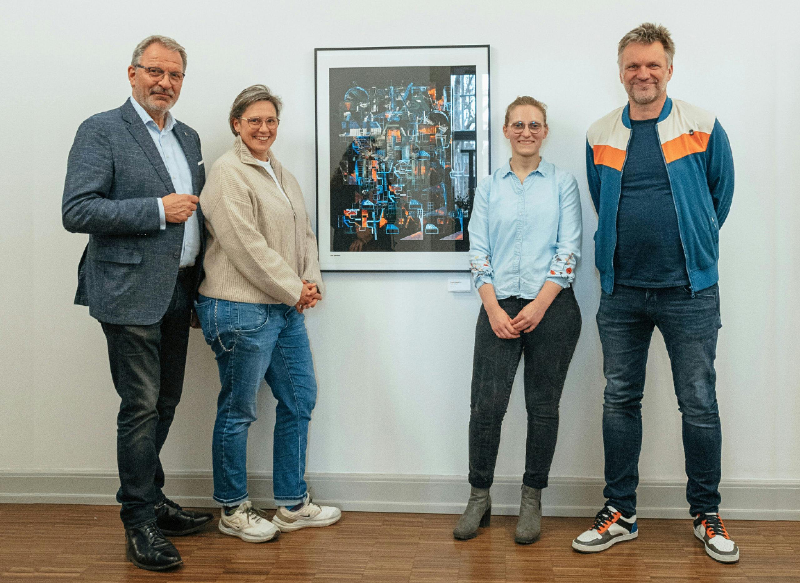 Fünf Personen stehen lächelnd nebeneinander vor einer weißen Wand in einer Ausstellung. In der Mitte hängt ein gerahmtes Kunstwerk, das aus leuchtenden, abstrakten Formen in Blau, Orange und Schwarz besteht. Der Boden ist mit Holzparkett ausgelegt. Die Gruppe wirkt entspannt und stolz, als würde sie das Kunstwerk gemeinsam präsentieren.