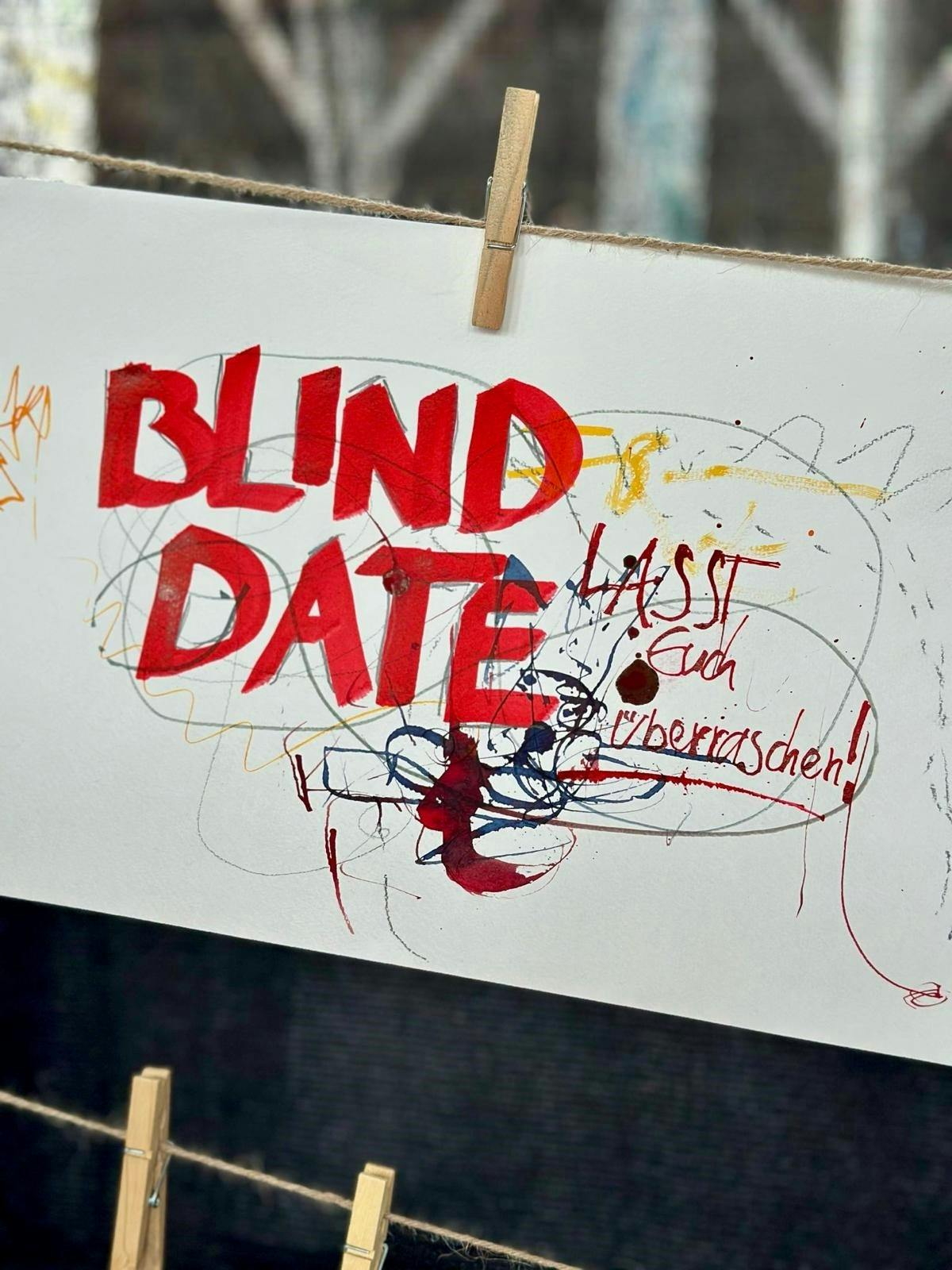 Ein weißes Blatt Papier hängt mit einer Holzklammer an einer Wäscheleine. In dicken roten Buchstaben steht darauf Blind Date geschrieben. Darunter und daneben ist mit dünneren Linien in verschiedenen Farben wie Gelb, Blau, Schwarz und Rot wild und abstrakt gekritzelt. In kleinerer roter Handschrift steht rechts daneben lässt euch überraschen. Das Blatt wirkt künstlerisch-experimentell gestaltet.