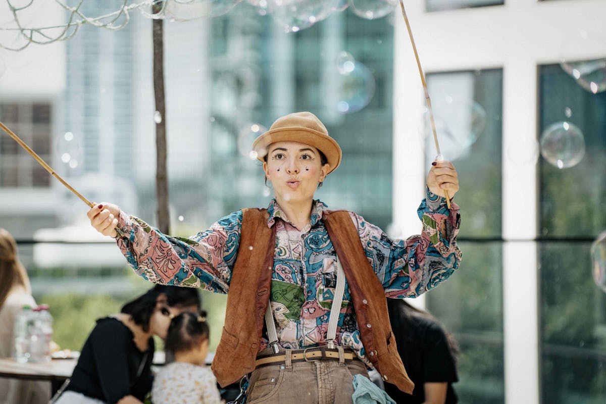 Eine Straßenkünstlerin steht im Mittelpunkt des Bildes und hält zwei Holzstäbe mit Seilen in den Händen, mit denen sie große Seifenblasen erzeugt. Sie trägt ein bunt gemustertes Hemd, eine braune Weste, Hosenträger und einen beigefarbenen Hut. Auf ihrem Gesicht sind kleine schwarze Punkte aufgemalt, die an ein Clowns-Make-up erinnern. Im Hintergrund sind verschwommen einige Erwachsene und Kinder zu sehen, die der Vorführung zusehen. Die Szene spielt sich in einem städtischen Umfeld mit modernen Gebäuden im Hintergrund ab. Die Stimmung ist fröhlich und verspielt.