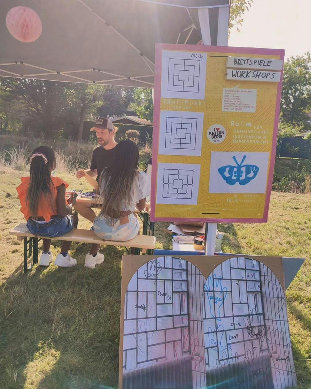 Ein Mann sitzt mit zwei Mädchen auf einer Holzbank unter einem Pavillon im Freien und erklärt ein Brettspiel. Daneben steht ein gelbes Plakat mit der Aufschrift Brettspiele Workshops und Zeichnungen des Spiels Mühle. Darunter befinden sich Logos und Informationen zum Projekt Katernberg. Im Vordergrund steht ein weiteres Plakat mit aufgemalten Händen und Namen. Die Szene spielt sich bei Sonnenschein auf einer Wiese ab.