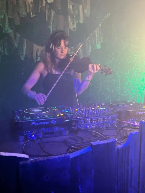 Eine Frau steht an einem DJ-Pult, trägt Kopfhörer und spielt gleichzeitig auf einer Geige. Das Pult ist mit Mischpulten und Plattenspielern der Marke Pioneer DJ ausgestattet. Die Szene ist in buntes Licht getaucht, mit einem bläulich-grünen Hintergrund und leichtem Nebel. Über dem DJ-Pult hängen dekorative Stoffstreifen. Die Atmosphäre wirkt kreativ und verbindet klassische Musik mit elektronischer Clubkultur.