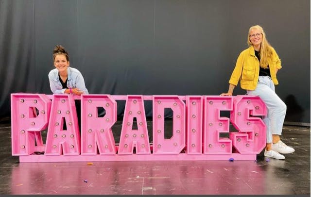 Zwei Frauen posieren lächelnd neben einer großen, pinken Leuchtschrift mit dem Wort Paradies. Die Buchstaben sind mit kleinen Glühbirnen besetzt. Die linke Frau lehnt sich mit dem Oberkörper auf die Buchstaben, die rechte sitzt lässig auf dem Buchstaben S. Der Hintergrund ist eine schlichte, dunkle Wand.