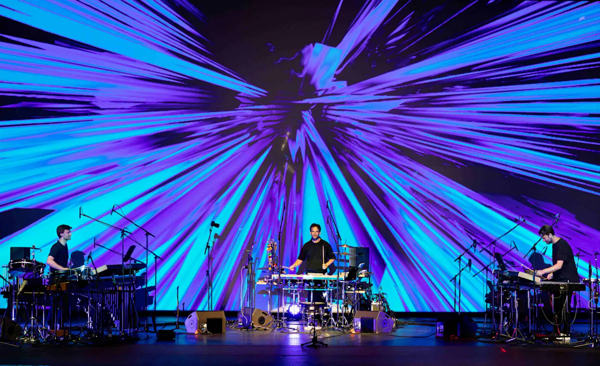 Drei Musiker stehen auf einer Bühne, umgeben von verschiedenen Percussion- und elektronischen Instrumenten. Im Hintergrund ist eine große Projektionsfläche zu sehen, auf der dynamische, strahlenförmige Lichtanimationen in Blau- und Lilatönen nach außen explodieren. Mehrere Mikrofone, Lautsprecher und Kabel sind über die Bühne verteilt. Die Szene wirkt technisch und visuell eindrucksvoll.