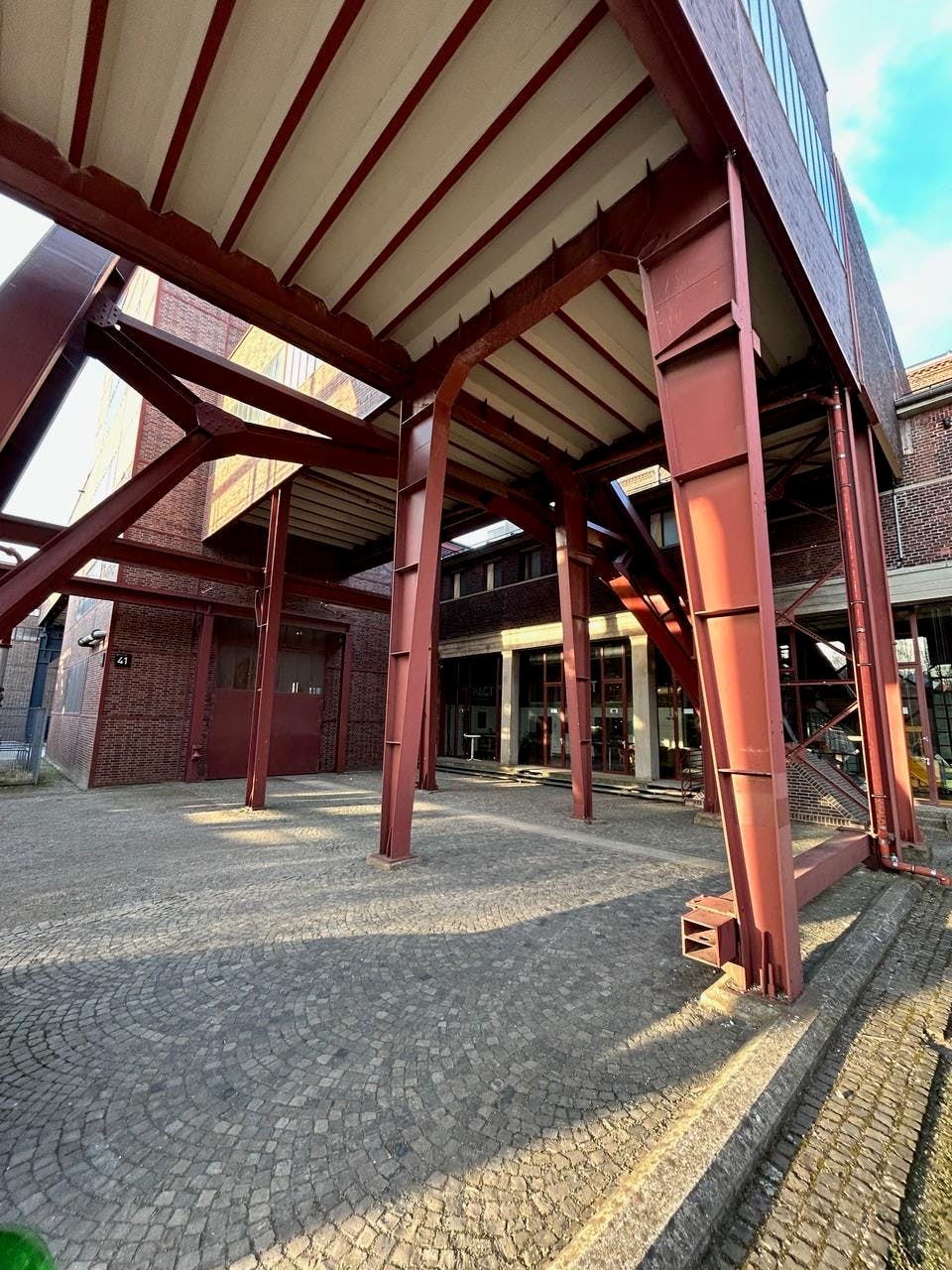 Blick von unten auf eine stählerne Tragstruktur auf dem Gelände der Zeche Zollverein in Essen, einem herausragenden Beispiel der Industriearchitektur der Neuen Sachlichkeit. Die roten Stahlträger und die klar gegliederte Backsteinfassade im Hintergrund betonen die funktionale Gestaltung ohne ornamentale Ausschmückung. Das Bild vermittelt das Prinzip der Sichtbarkeit von Konstruktion und Zweck, das typisch für die sachlich-nüchterne Architektur der 1920er Jahre ist. Die gepflasterte Fläche im Vordergrund ergänzt den industriellen Charakter des Ensembles.