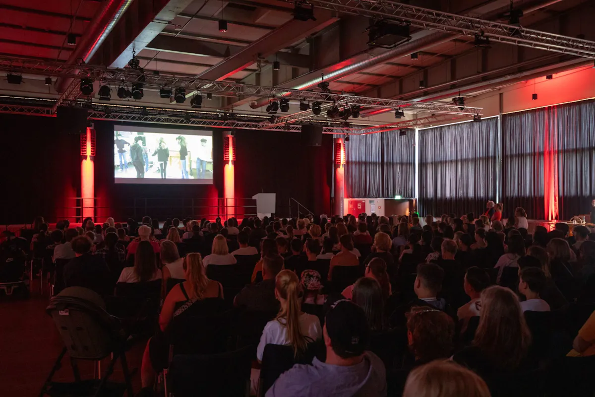 Eine große Menschenmenge sitzt in einem abgedunkelten Veranstaltungssaal und blickt auf eine Leinwand, auf der ein Film oder eine Präsentation gezeigt wird. Der Raum ist mit rotem Licht stimmungsvoll ausgeleuchtet. An der Decke sind zahlreiche Scheinwerfer und Technikträger installiert. Die Fenster an der Seite sind mit dunklen Vorhängen abgedeckt. Das Publikum wirkt aufmerksam und ruhig.