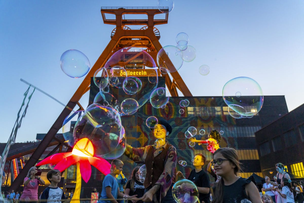 Menschen feiern vor dem markanten Förderturm der Zeche Zollverein bei einem Open-Air-Event in der Abenddämmerung. Über ihnen schweben zahlreiche große Seifenblasen. Im Vordergrund steht eine Künstlerin mit Hut und bunter Kleidung, der Seifenblasen macht. Kinder und Erwachsene schauen begeistert zu, während bunte Lichtinstallationen und eine leuchtende Blumenfigur die Szenerie ergänzen. Die Atmosphäre ist lebendig und verspielt.