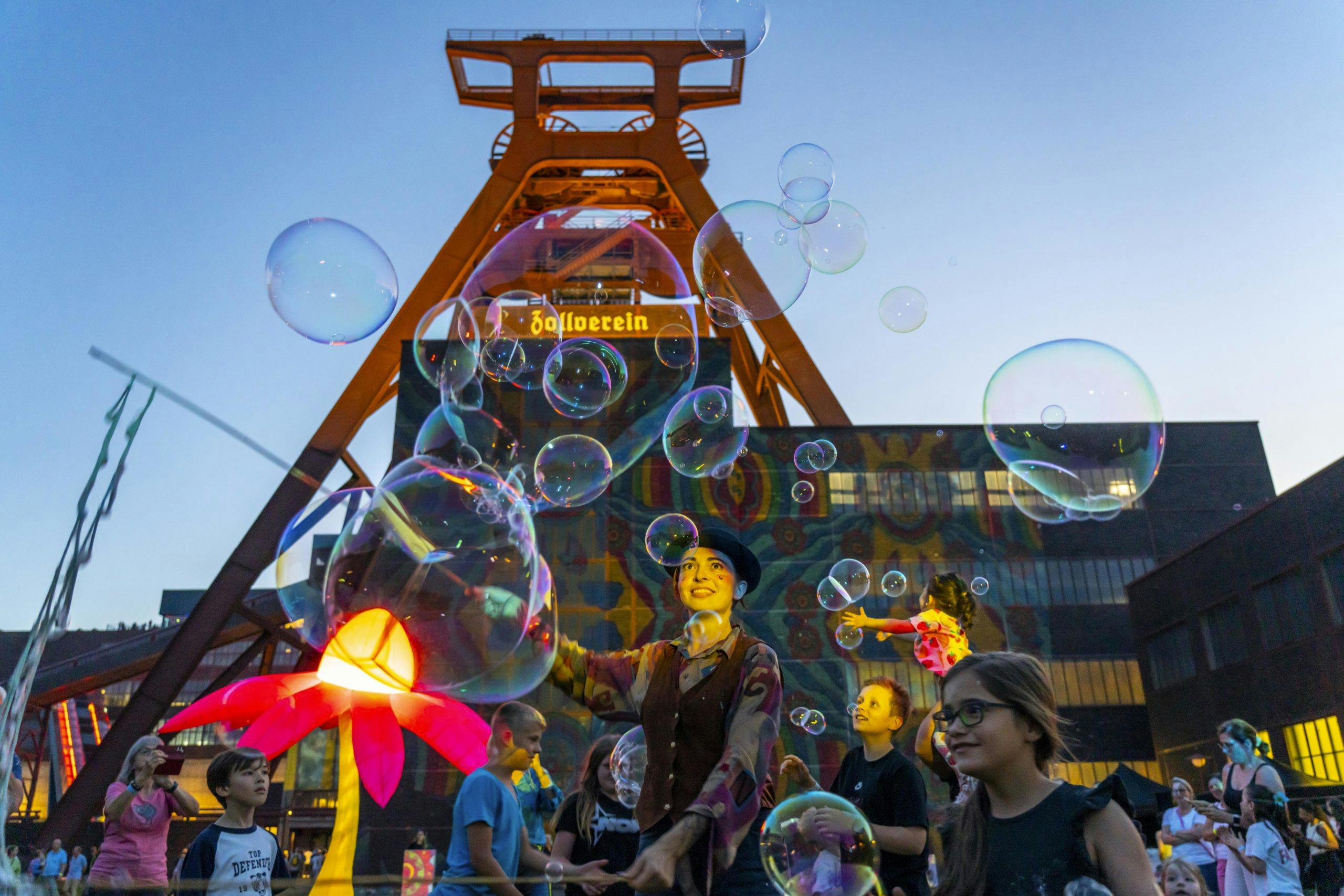 Menschen feiern vor dem markanten Förderturm der Zeche Zollverein bei einem Open-Air-Event in der Abenddämmerung. Über ihnen schweben zahlreiche große Seifenblasen. Im Vordergrund steht eine Künstlerin mit Hut und bunter Kleidung, der Seifenblasen macht. Kinder und Erwachsene schauen begeistert zu, während bunte Lichtinstallationen und eine leuchtende Blumenfigur die Szenerie ergänzen. Die Atmosphäre ist lebendig und verspielt.