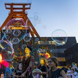 Menschen feiern vor dem markanten Förderturm der Zeche Zollverein bei einem Open-Air-Event in der Abenddämmerung. Über ihnen schweben zahlreiche große Seifenblasen. Im Vordergrund steht eine Künstlerin mit Hut und bunter Kleidung, der Seifenblasen macht. Kinder und Erwachsene schauen begeistert zu, während bunte Lichtinstallationen und eine leuchtende Blumenfigur die Szenerie ergänzen. Die Atmosphäre ist lebendig und verspielt.