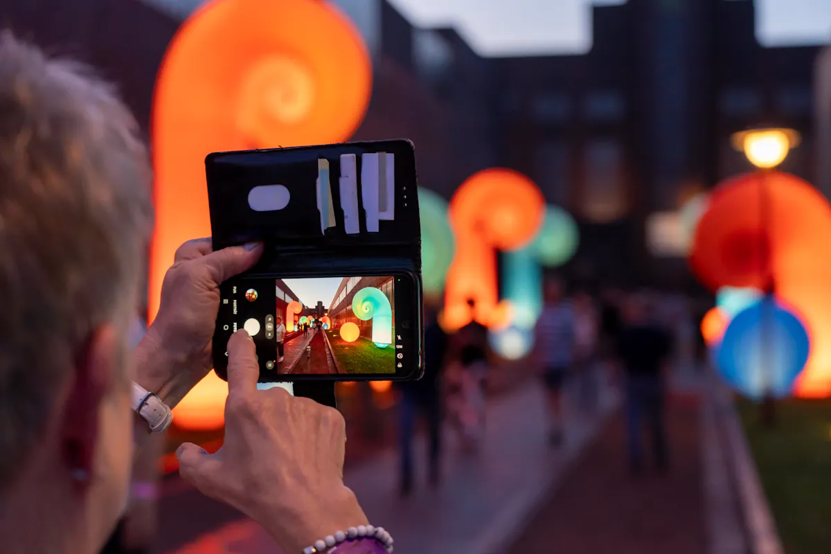 Eine Person hält ein Smartphone in einer schwarzen Hülle und fotografiert eine nächtliche Straßenszene mit leuchtenden, bunten Spiralskulpturen. Auf dem Handybildschirm ist die Szene mit den Kunstobjekten und vorbeigehenden Menschen scharf zu erkennen, während der Hintergrund des Fotos verschwommen ist. Die Szenerie ist farbenfroh und lebendig beleuchtet.