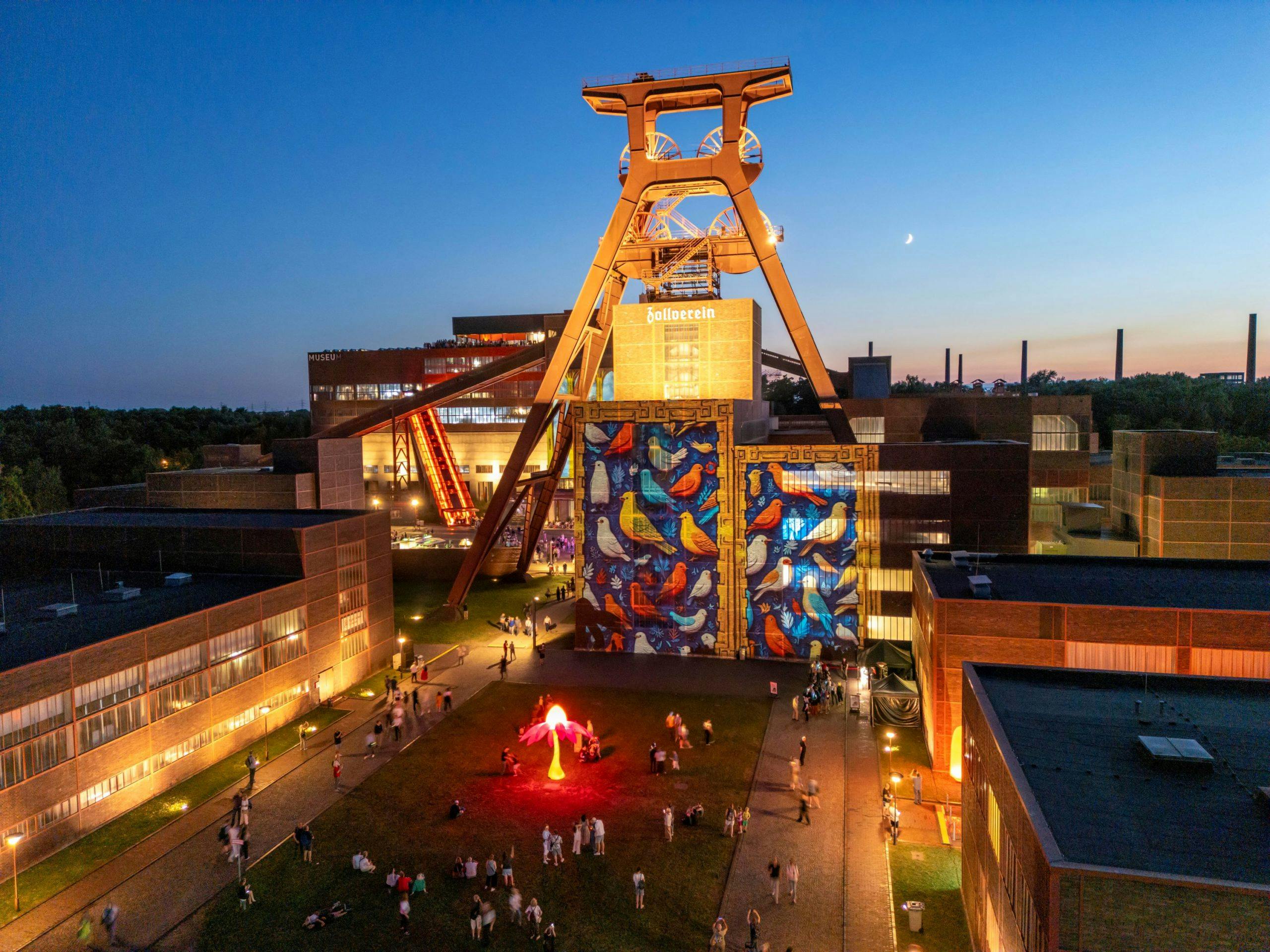 Abendaufnahme des UNESCO-Welterbes Zeche Zollverein in Essen. Im Mittelpunkt steht das beleuchtete Doppelbock-Fördergerüst mit der Aufschrift Zollverein. Die Fassade des Gebäudes darunter ist mit einem großformatigen bunten Kunstwerk mit Vogelmotiven gestaltet. Im Vordergrund befindet sich ein beleuchtetes, pink leuchtendes Objekt in Form einer Blume, umgeben von spazierenden und verweilenden Menschen. Die umliegenden Gebäude der ehemaligen Zeche sind ebenfalls dezent beleuchtet, während im Hintergrund die untergehende Sonne den Himmel in blau-orange färbt.