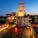 Abendaufnahme des UNESCO-Welterbes Zeche Zollverein in Essen. Im Mittelpunkt steht das beleuchtete Doppelbock-Fördergerüst mit der Aufschrift Zollverein. Die Fassade des Gebäudes darunter ist mit einem großformatigen bunten Kunstwerk mit Vogelmotiven gestaltet. Im Vordergrund befindet sich ein beleuchtetes, pink leuchtendes Objekt in Form einer Blume, umgeben von spazierenden und verweilenden Menschen. Die umliegenden Gebäude der ehemaligen Zeche sind ebenfalls dezent beleuchtet, während im Hintergrund die untergehende Sonne den Himmel in blau-orange färbt.