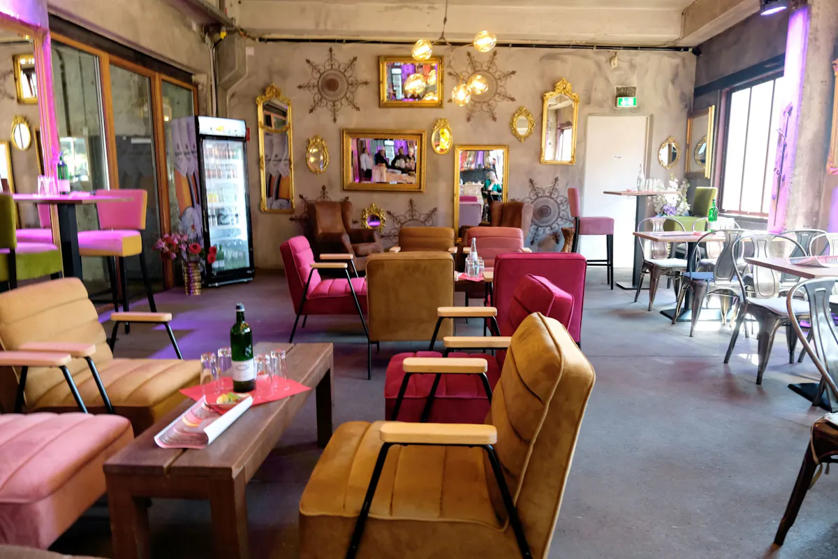 Gemütlich eingerichtetes Café oder Lounge mit einer Mischung aus modernen und vintage-inspirierten Möbeln. Verschiedene bunte Polstersessel in Rosa-, Braun- und Senftönen stehen um niedrige Holztische, auf denen Gläser und Flaschen stehen. Im Hintergrund hängen goldgerahmte Spiegel und verzierte Wanddekorationen an einer grauen Betonwand. Links steht ein Kühlschrank mit Getränken, rechts sind Tische mit Stühlen für Speisende gedeckt. Das Licht ist warm, mit dezenten pinken und violetten Akzenten, was eine einladende Atmosphäre schafft.