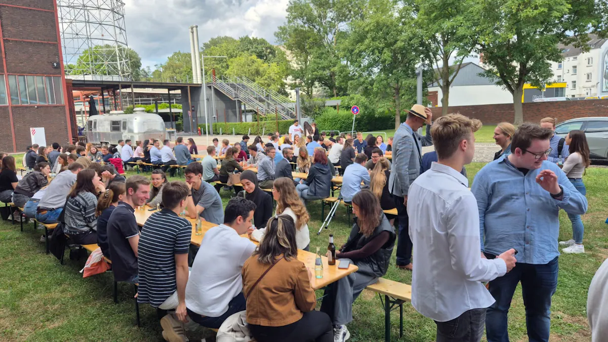 Dutzende Menschen sitzen an langen Holzbänken auf einer Wiese vor einem alten Industriegebäude und unterhalten sich lebhaft. Die Stimmung ist ausgelassen und freundlich. Im Hintergrund sind ein silberner Wohnwagen, ein Stahlgitterturm und eine breite Metalltreppe zu sehen. Die Szenerie wirkt wie ein gemeinsames Sommerfest oder ein informelles Treffen bei angenehmem Wetter.