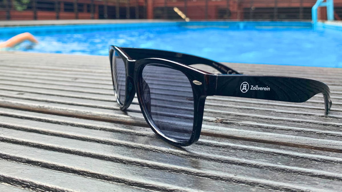 Eine schwarze Sonnenbrille mit dem Logo und Schriftzug Zollverein liegt auf einem hölzernen Pooldeck. Im Hintergrund ist ein blaues Schwimmbecken zu sehen, in dem eine Person schwimmt. Die Szene wirkt sommerlich und entspannt.
