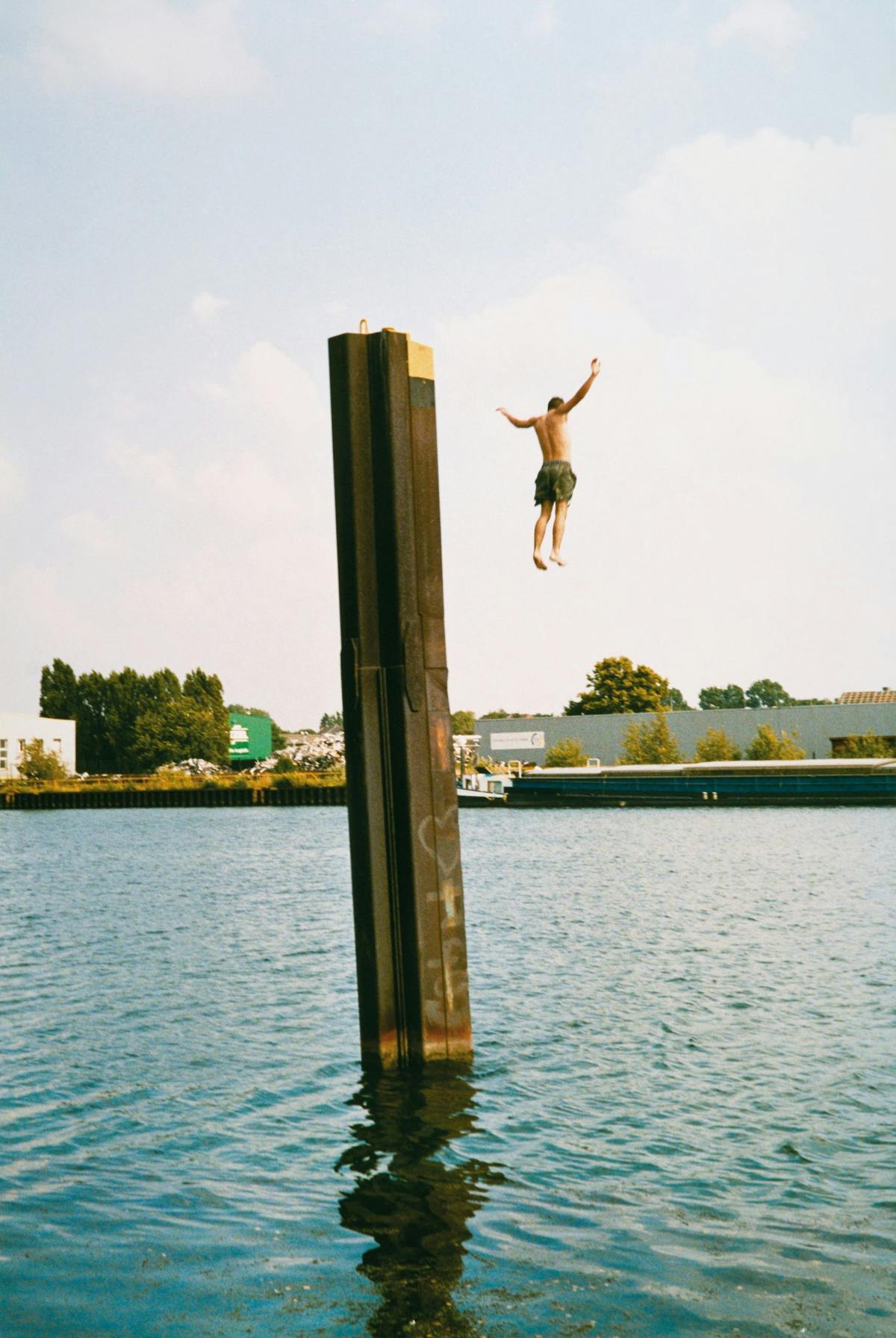 Ein Mann springt mit ausgebreiteten Armen von einem hohen Stahlpfeiler in ein Gewässer, unter ihm spiegelt sich der Pfeiler im Wasser, im Hintergrund sind Industriehallen, Bäume und ein Frachtschiff zu sehen.