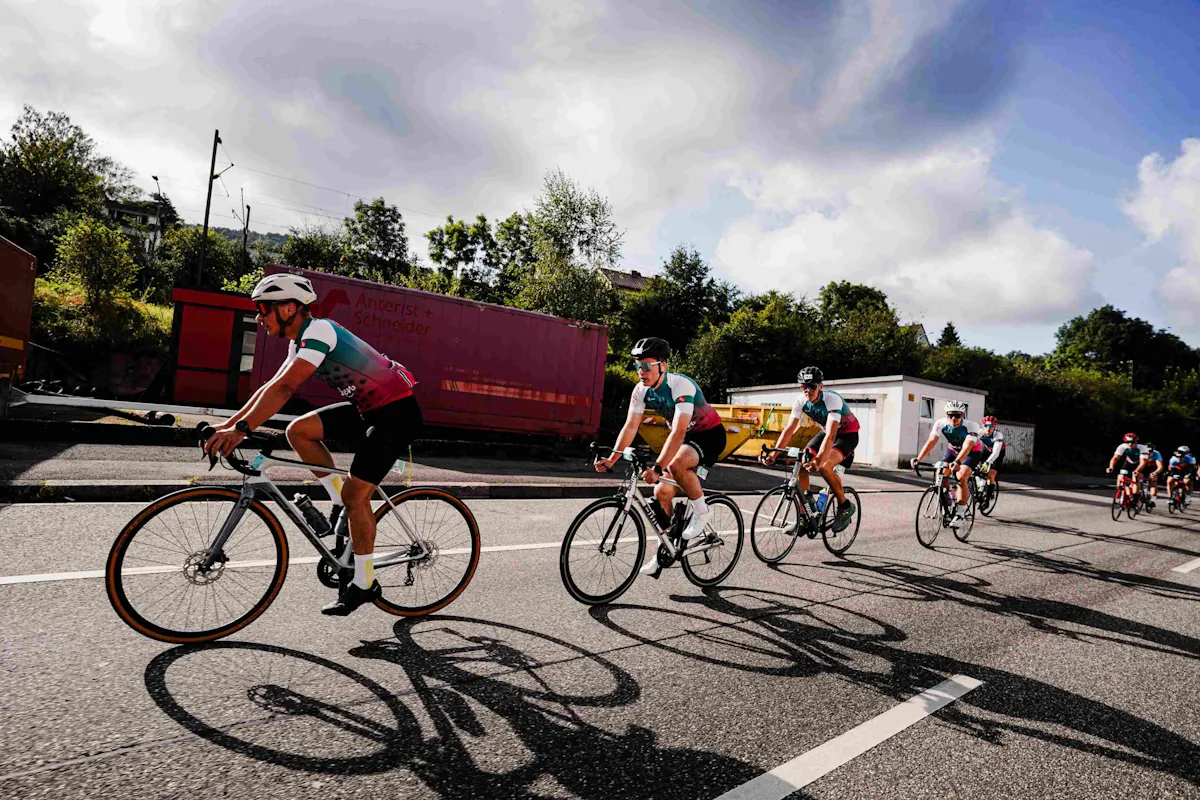 Das Bild zeigt eine Gruppe von Radfahrern, die in sportlicher Kleidung und Helmen auf einer Straße unterwegs sind. Sie fahren in einer Reihe dicht hintereinander, einige schauen konzentriert nach vorne. Die Räder werfen deutliche Schatten auf den Asphalt. Im Hintergrund sind Bäume, ein weißes Gebäude und rote Container zu sehen, über denen sich ein teils bewölkter, teils blauer Himmel ausbreitet.