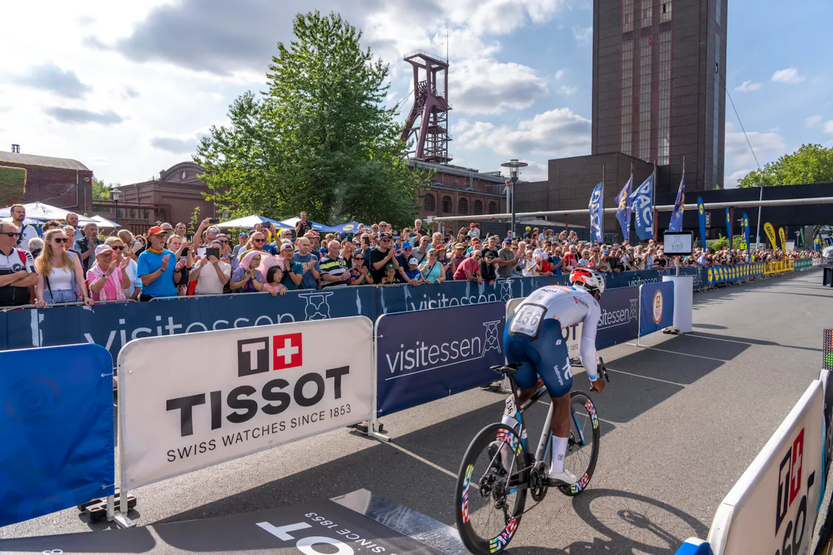 Ein Radrennfahrer mit Startnummer 136 fährt an jubelnden Zuschauern vorbei, im Hintergrund sind Banner, Absperrgitter und das Fördergerüst der Zeche Zollverein zu sehen.
