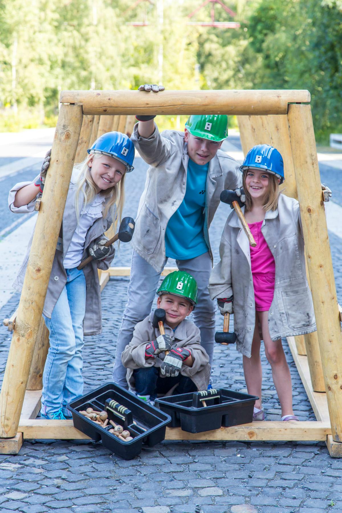 Vier Kinder tragen Helme und Arbeitsjacken und spielen im Freien mit Holzgerüsten und Werkzeugen. Zwei Kinder halten Hämmer in den Händen, während zwei Werkzeugkästen mit Holzutensilien vor ihnen stehen. Alle lachen und wirken fröhlich, im Hintergrund sind Bäume und Kopfsteinpflaster zu sehen.