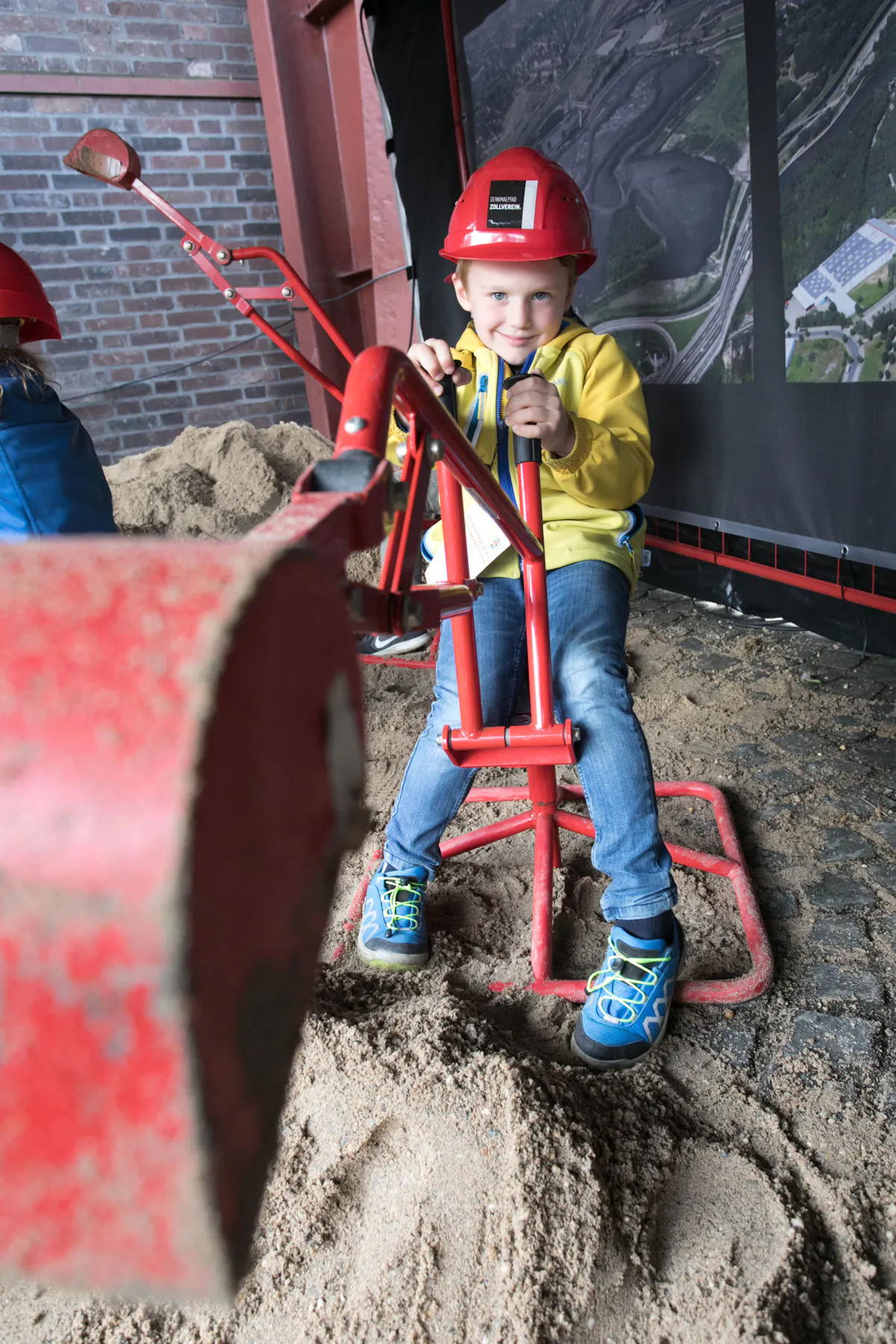 Ein Kind mit rotem Schutzhelm und gelber Jacke sitzt auf einem kleinen Bagger-Spielgerät und gräbt im Sand. Es hält die Steuerhebel fest und lächelt in die Kamera. Im Hintergrund sind eine Ziegelwand und ein großes Foto einer Industrieanlage zu sehen.