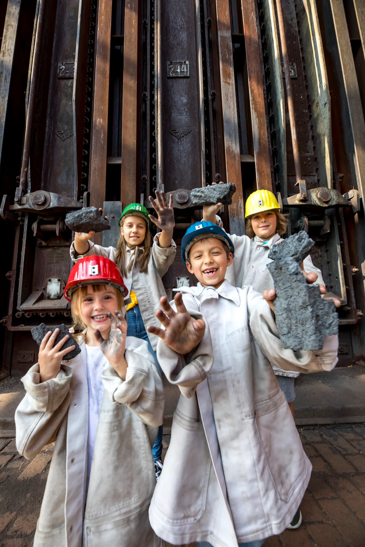 Vier Kinder tragen bunte Schutzhelme und helle Arbeitsjacken. Sie stehen lachend vor einer großen rostigen Industrieanlage und halten Stücke von dunkler Kohle in den Händen. Ihre Gesichter und Hände sind vom Staub verschmutzt, was die Szene spielerisch und lebendig wirken lässt.