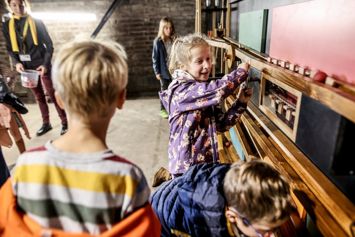 Mehrere Kinder spielen und experimentieren an einer interaktiven Holzinstallation mit bunten Elementen in einem Raum mit Backsteinwänden. Ein Mädchen in einer lilafarbenen Jacke dreht lachend an einer Kurbel, während andere Kinder neugierig zusehen. Im Hintergrund steht eine Aufsichtsperson mit Namensschild.