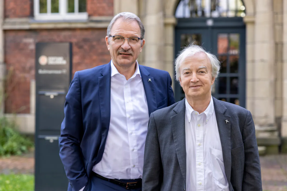 Zwei Männer stehen nebeneinander vor dem Eingang eines historischen Gebäudes mit hellem Sandsteinportal und dunklen Türen mit Glasfenstern. Der Mann links trägt einen dunkelblauen Anzug, ein weißes Hemd und eine Brille mit dunklem Rand. Er hat graue Haare und einen Schnurrbart. Der Mann rechts trägt einen grauen Anzug mit weißem Hemd, hat graue, leicht zerzauste Haare und lächelt freundlich. Beide tragen am Revers ihres Sakkos eine kleine Anstecknadel in Form des Fördergerüsts der Zeche Zollverein. Im Hintergrund ist ein Schild mit der Aufschrift Stiftung Zollverein zu sehen.