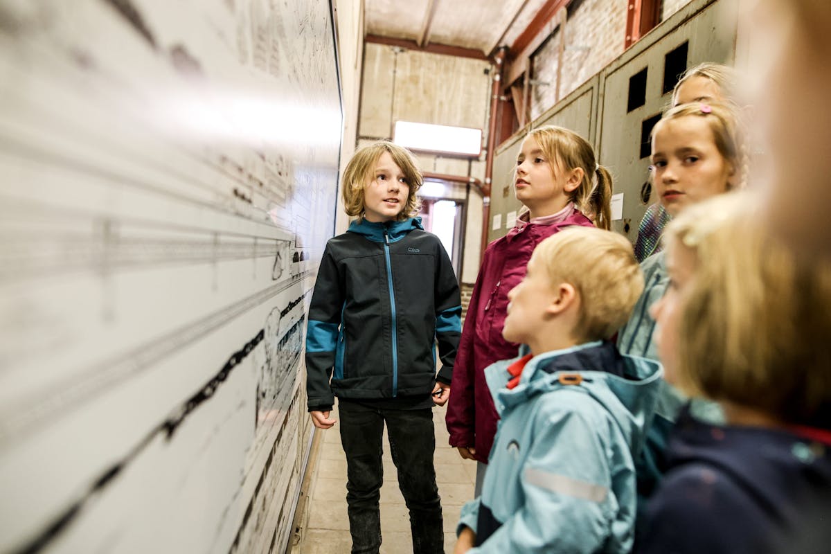 Eine Gruppe von Kindern steht in einer Industriehalle und betrachtet aufmerksam eine große Wandtafel mit technischen Zeichnungen. Die Kinder tragen wetterfeste Jacken und wirken interessiert und neugierig. Im Hintergrund sind alte Metallkonstruktionen und Wände aus Backstein zu sehen.