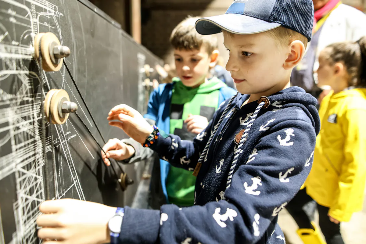 Ein Junge mit dunkelblauer Kapuzenjacke und Baseballkappe bedient konzentriert eine Wandinstallation mit Seilen und Rollen, die technische Abläufe veranschaulicht. Neben ihm steht ein weiterer Junge und beobachtet interessiert. Im Hintergrund sind weitere Kinder in bunter Kleidung zu sehen.