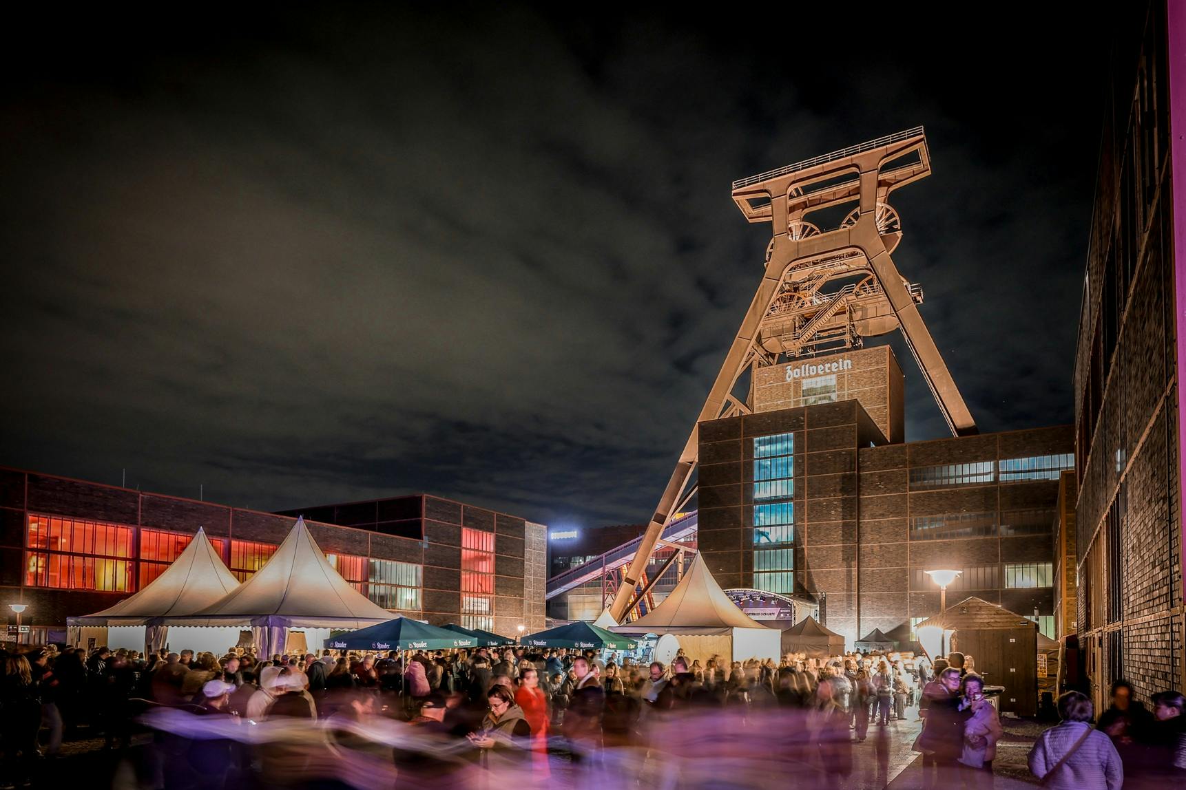 Abendszene auf dem Gelände der Zeche Zollverein mit dem beleuchteten Förderturm im Hintergrund. Viele Menschen bewegen sich zwischen Zelten und Ständen, die in warmes Licht getaucht sind. Die historischen Backsteingebäude leuchten in verschiedenen Farben und schaffen eine festliche Atmosphäre unter einem wolkigen Nachthimmel.