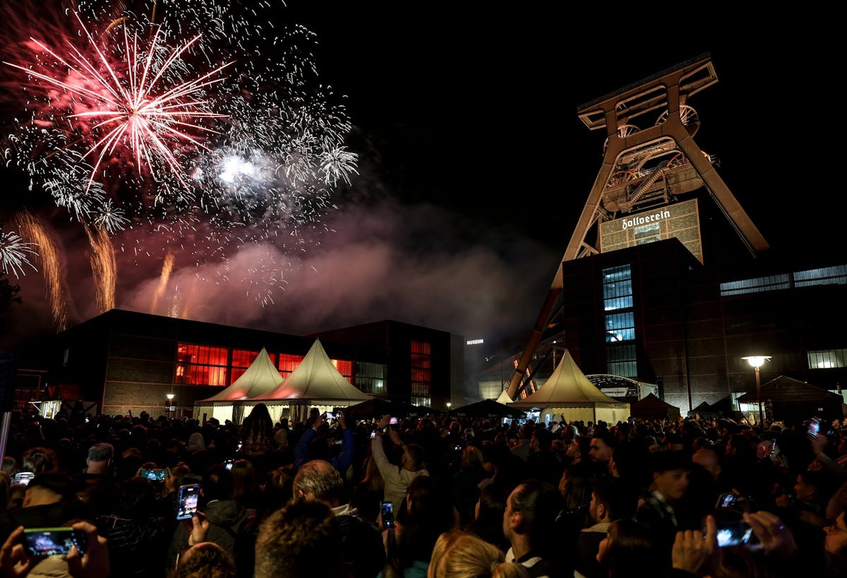 Ein großes Feuerwerk mit roten, weißen und goldenen Lichtern erhellt den Nachthimmel über dem Förderturm der Zeche Zollverein. Vor den hell erleuchteten Backsteingebäuden und weißen Festzelten steht eine dichte Menschenmenge, viele halten ihre Handys hoch, um das Spektakel zu fotografieren oder zu filmen.