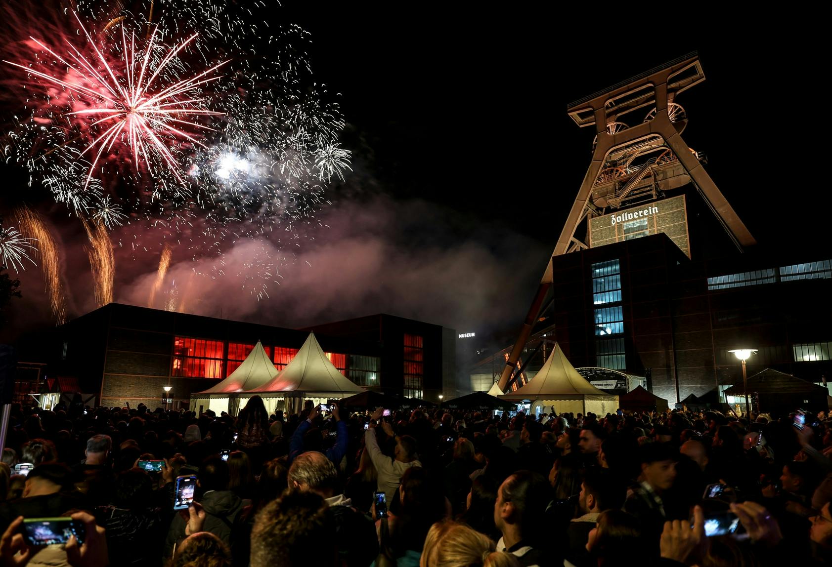Ein großes Feuerwerk mit roten, weißen und goldenen Lichtern erhellt den Nachthimmel über dem Förderturm der Zeche Zollverein. Vor den hell erleuchteten Backsteingebäuden und weißen Festzelten steht eine dichte Menschenmenge, viele halten ihre Handys hoch, um das Spektakel zu fotografieren oder zu filmen.