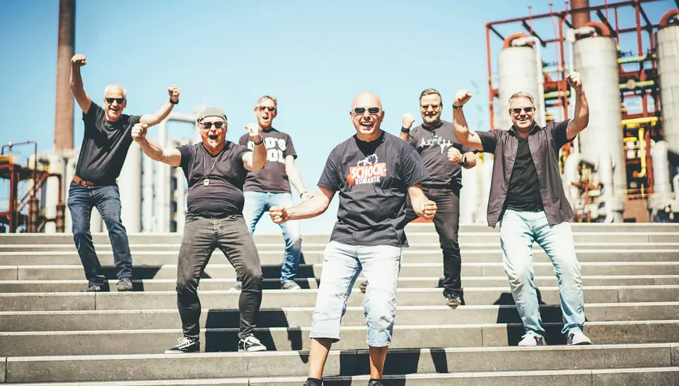 Sechs Männer posieren lachend auf einer breiten Treppe im Freien. Alle tragen lässige schwarze T-Shirts und Sonnenbrillen. Der Mann in der Mitte vorne hat kurze Haare, trägt eine helle kurze Jeanshose und ein schwarzes T-Shirt mit rotem Aufdruck. Die Männer zeigen energische Posen mit erhobenen Fäusten. Im Hintergrund sind Industrieanlagen mit Rohren und Türmen zu sehen, darüber ein blauer Himmel.