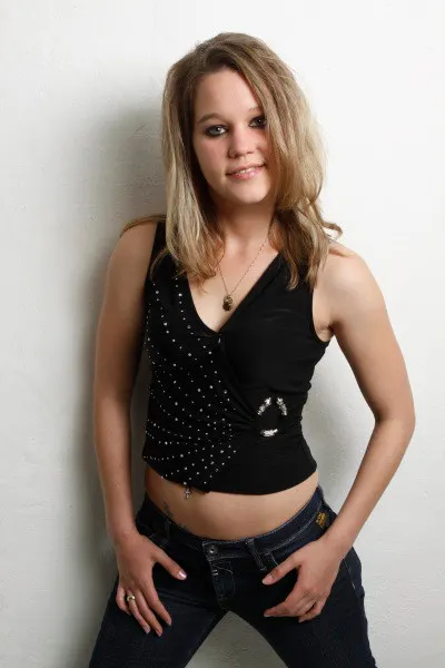 Eine junge Frau mit schulterlangen blonden Haaren steht vor einer weißen Wand und blickt lächelnd in die Kamera. Sie trägt ein schwarzes ärmelloses Top mit Glitzersteinen und eine dunkle Jeans. Ihre Hände liegen locker in den Hosentaschen, und sie trägt eine Halskette mit Anhänger.