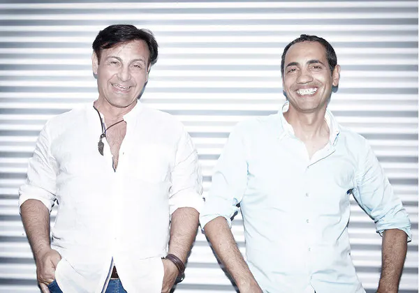 Zwei Männer stehen nebeneinander und lächeln in die Kamera. Beide tragen helle Hemden, der Mann links ein weißes Hemd mit aufgeknöpftem Kragen und einem auffälligen Anhänger an einer Kette, der Mann rechts ein hellblaues Hemd mit hochgekrempelten Ärmeln. Beide haben die Hände in den Hosentaschen. Der Hintergrund besteht aus einer Wand mit horizontalen Streifen.