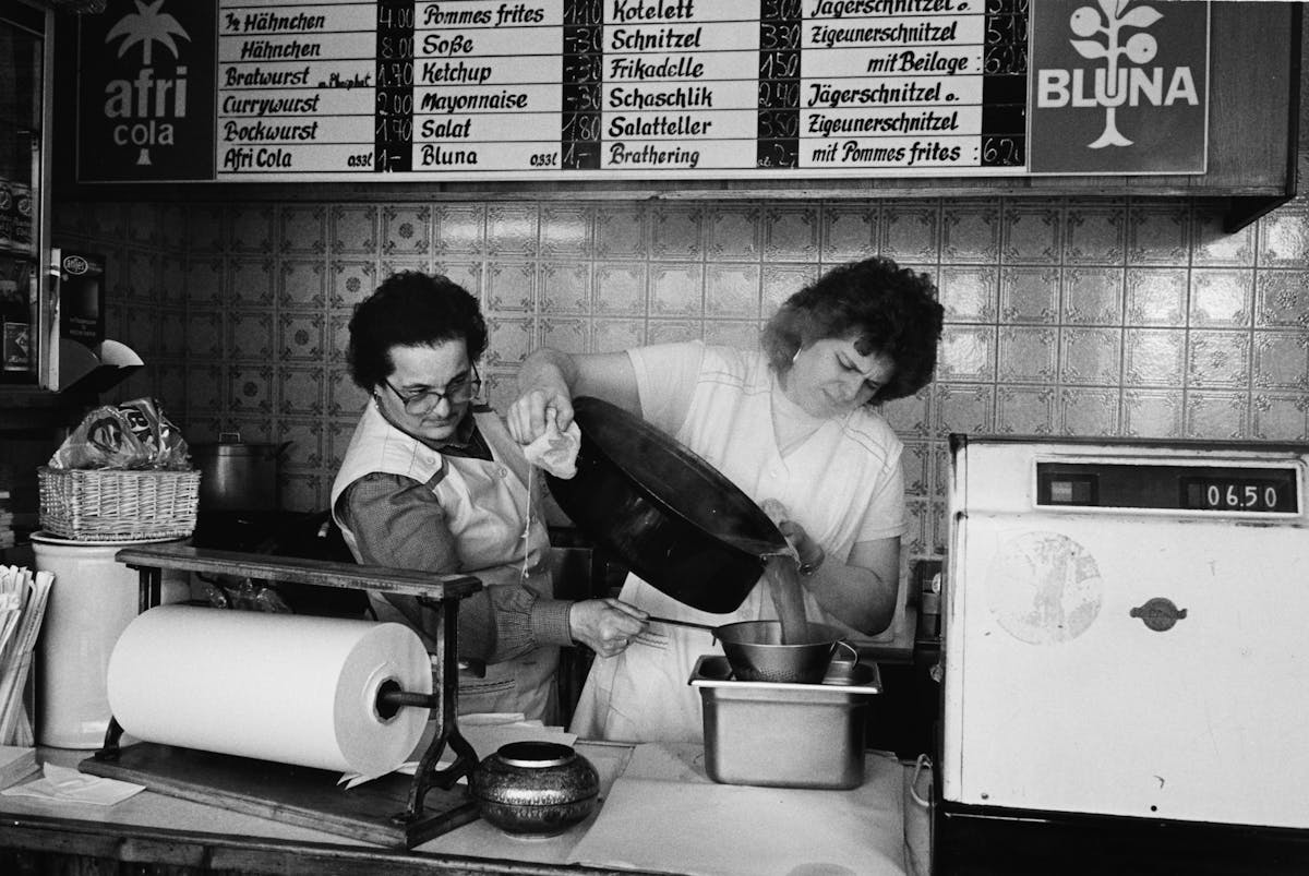Schwarzweißfoto aus einer Imbissbude, in der zwei Frauen in weißen Schürzen arbeiten. Eine gießt Suppe oder Soße aus einem großen Topf durch ein Sieb, die andere hält das Gefäß darunter. Im Hintergrund hängen alte Speisetafeln mit Preisen für Gerichte wie Schnitzel, Currywurst, Pommes frites und Jägerschnitzel. An den Wänden sind Werbeschilder für Afri-Cola und Bluna zu sehen.