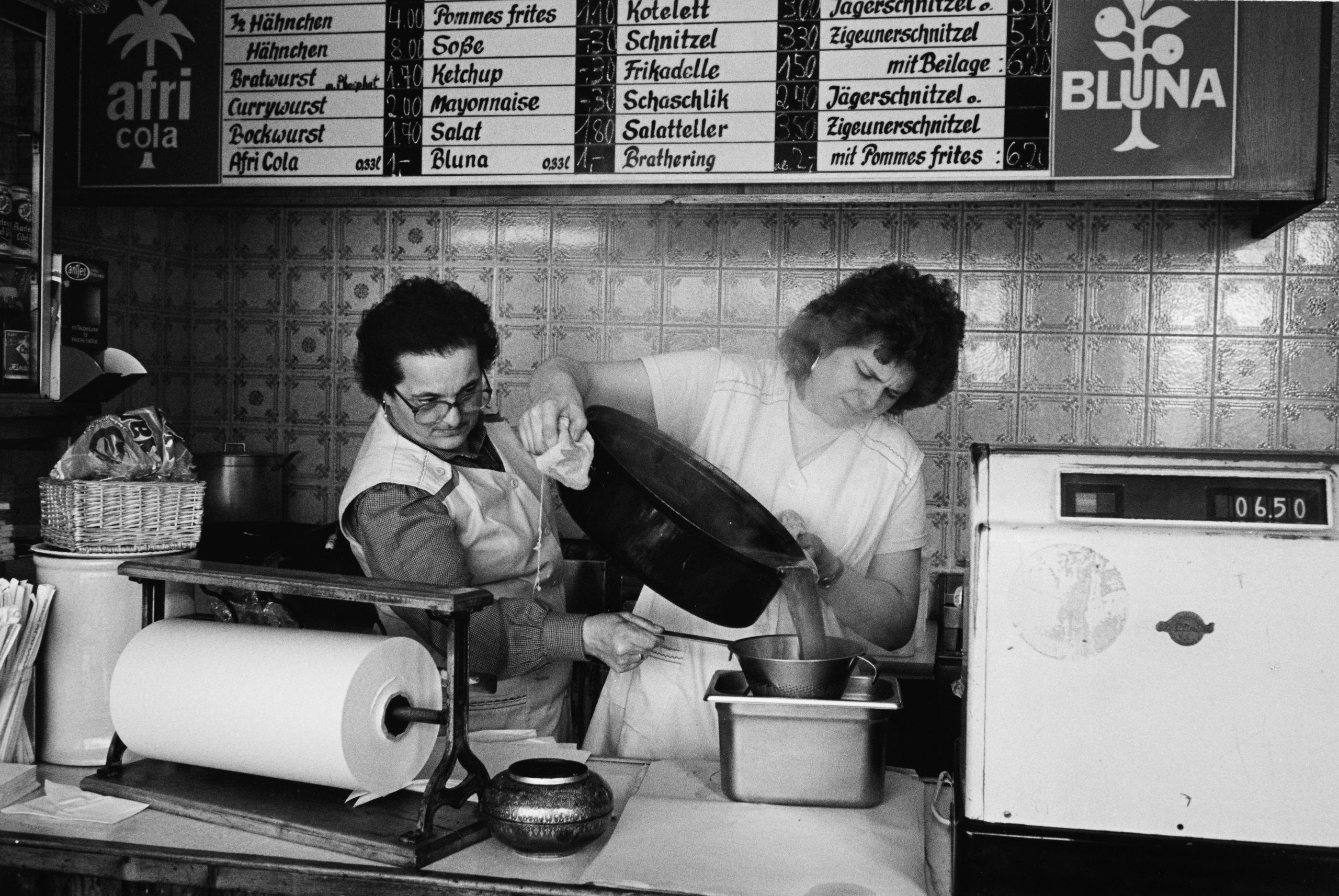 Schwarzweißfoto aus einer Imbissbude, in der zwei Frauen in weißen Schürzen arbeiten. Eine gießt Suppe oder Soße aus einem großen Topf durch ein Sieb, die andere hält das Gefäß darunter. Im Hintergrund hängen alte Speisetafeln mit Preisen für Gerichte wie Schnitzel, Currywurst, Pommes frites und Jägerschnitzel. An den Wänden sind Werbeschilder für Afri-Cola und Bluna zu sehen.