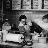 Schwarzweißfoto aus einer Imbissbude, in der zwei Frauen in weißen Schürzen arbeiten. Eine gießt Suppe oder Soße aus einem großen Topf durch ein Sieb, die andere hält das Gefäß darunter. Im Hintergrund hängen alte Speisetafeln mit Preisen für Gerichte wie Schnitzel, Currywurst, Pommes frites und Jägerschnitzel. An den Wänden sind Werbeschilder für Afri-Cola und Bluna zu sehen.