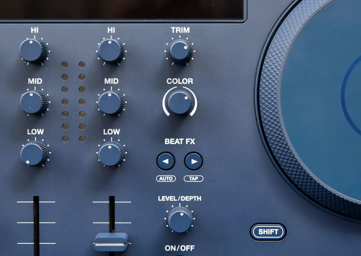 Nahaufnahme eines DJ-Mischpults mit mehreren Drehknöpfen, Schiebereglern und Tasten. Beschriftungen wie HI, MID, LOW, COLOR und BEAT FX sind sichtbar, ebenso eine große Jog-Wheel-Scheibe auf der rechten Seite. Das Gerät ist in dunklem Blau gehalten.