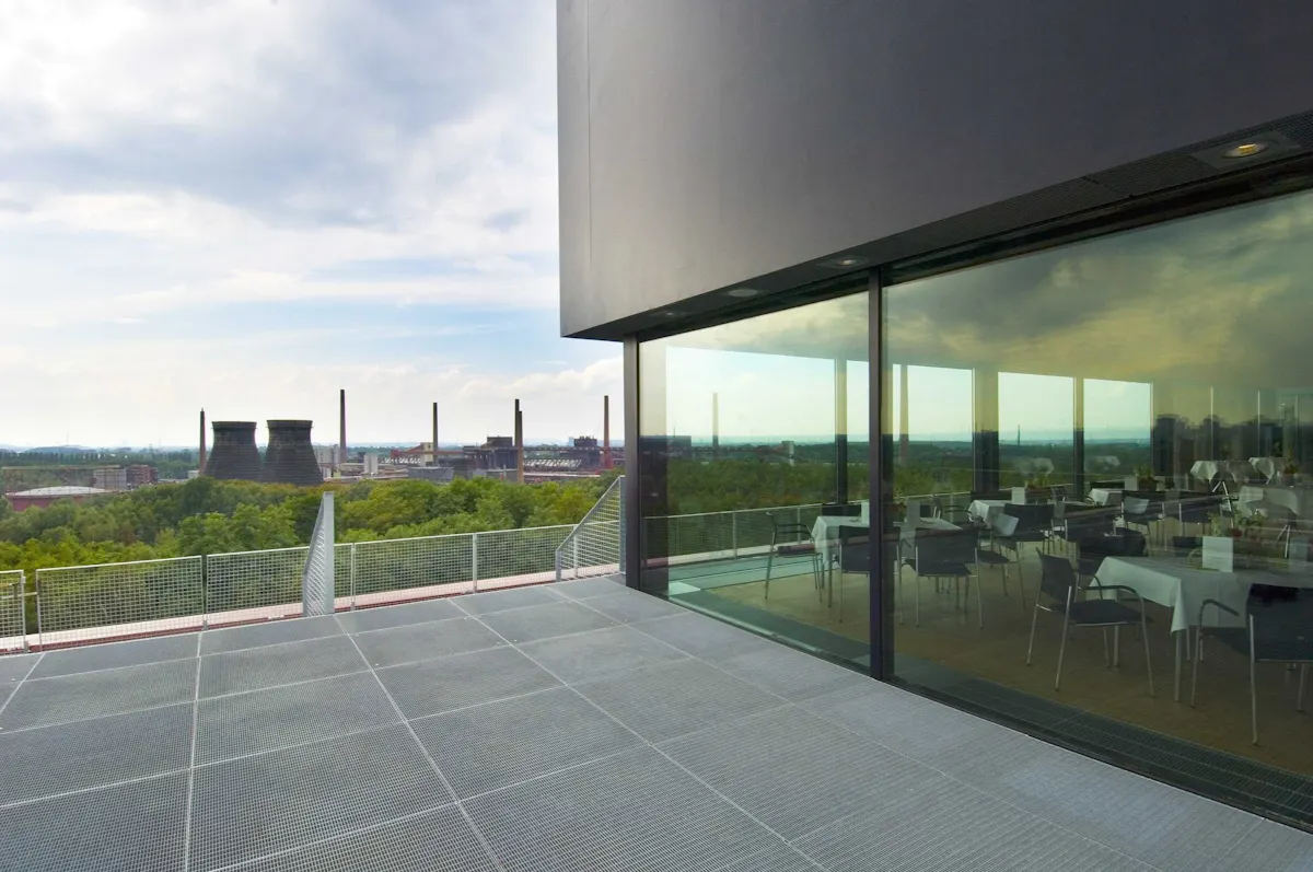 Eine moderne Terrasse mit Metallboden und Glasgeländer bietet einen weiten Blick über ein grünes Waldgebiet und industrielle Gebäude mit Schornsteinen und Kühltürmen in der Ferne. Rechts spiegelt sich die Landschaft in den großen Fenstern eines Saals mit gedeckten Tischen.