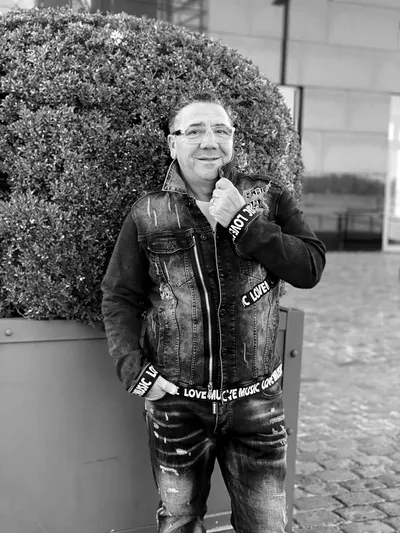 Ein Mann steht lächelnd vor einem großen runden Strauch in einem Pflanzkübel. Er trägt eine Jeansjacke mit auffälligen Schriftzügen, darunter ein schwarzes Oberteil und eine Jeanshose mit Used-Look. Eine Hand steckt in der Hosentasche, mit der anderen fasst er an den Kragen seiner Jacke. Das Bild ist in Schwarz-Weiß aufgenommen.